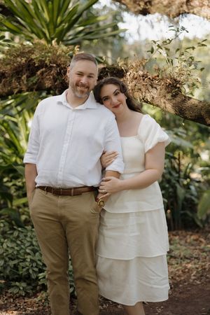 A romantic couple stands close together in a tropical garden setting wearing white and khaki clothing.