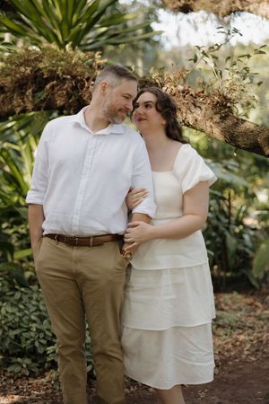 An intimate moment between a couple in white and khaki attire standing among tropical plants and trees.