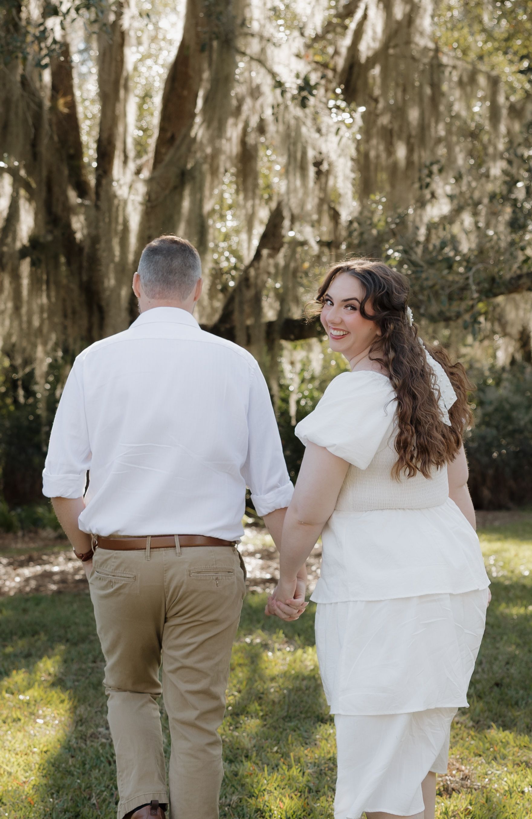 A couple in white outfits holding hands while walking through a garden with Spanish moss hanging from trees.