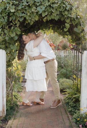A couple embraces in a romantic moment under lush green foliage along a garden pathway with white pillars.