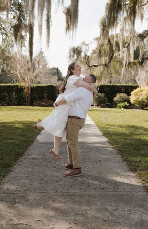 A couple in white and khaki outfits share a romantic dance move on a path beneath Spanish moss draped trees.