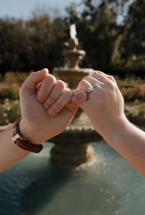 Two hands link pinky fingers in a promise gesture with an engagement ring visible and a fountain in the background.