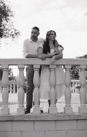 A couple leans on a white balustrade railing while enjoying a romantic outdoor moment in black and white photography.