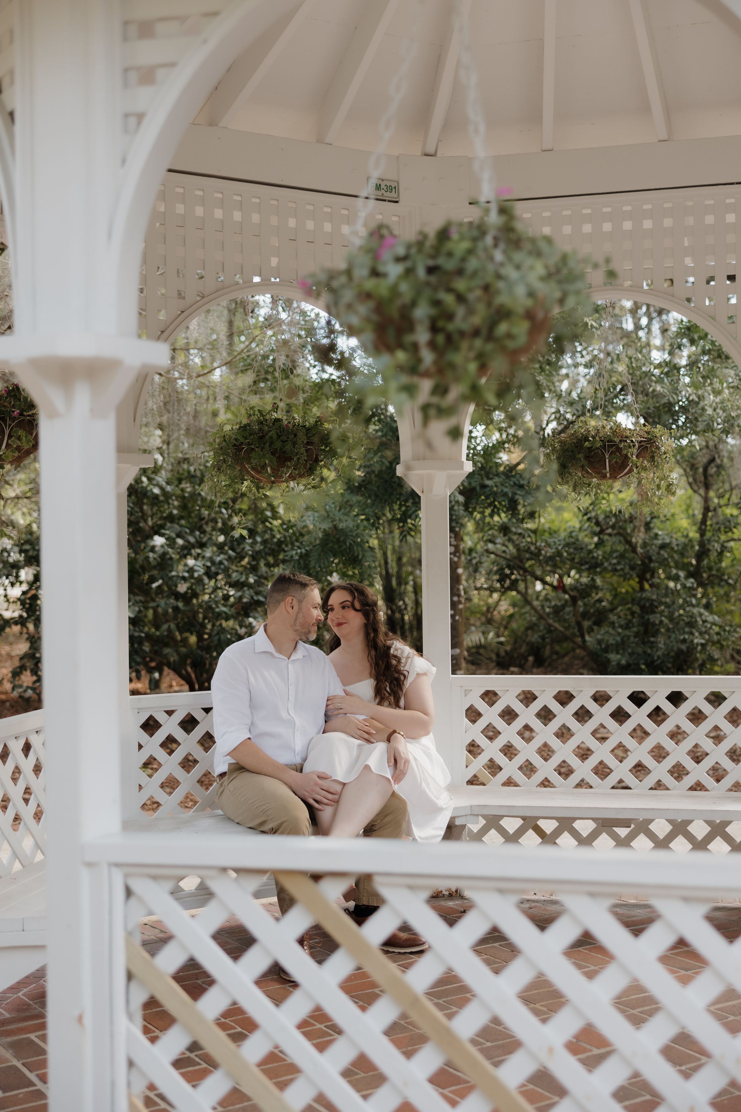 A romantic couple shares tender moments in a white latticed gazebo surrounded by lush greenery and string lights.
