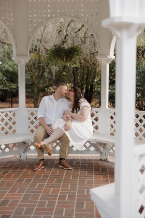 A couple sits together on a white latticed porch bench during a romantic outdoor moment.