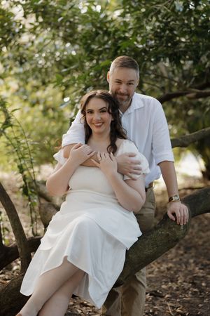 A romantic outdoor photoshoot of a couple in white clothing sitting on a fallen tree branch in a forest setting.