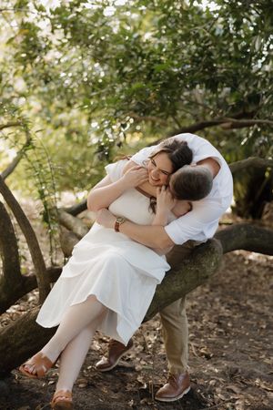 A couple in white clothing shares intimate moments while sitting on a tree branch in a romantic outdoor setting.
