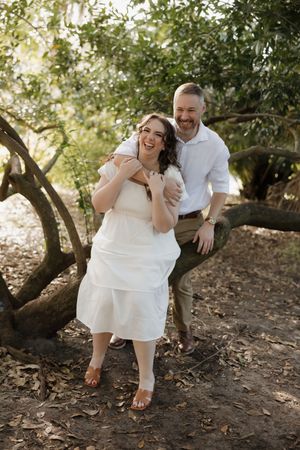 A couple in white and khaki outfits share joyful candid moments together in a wooded outdoor setting.