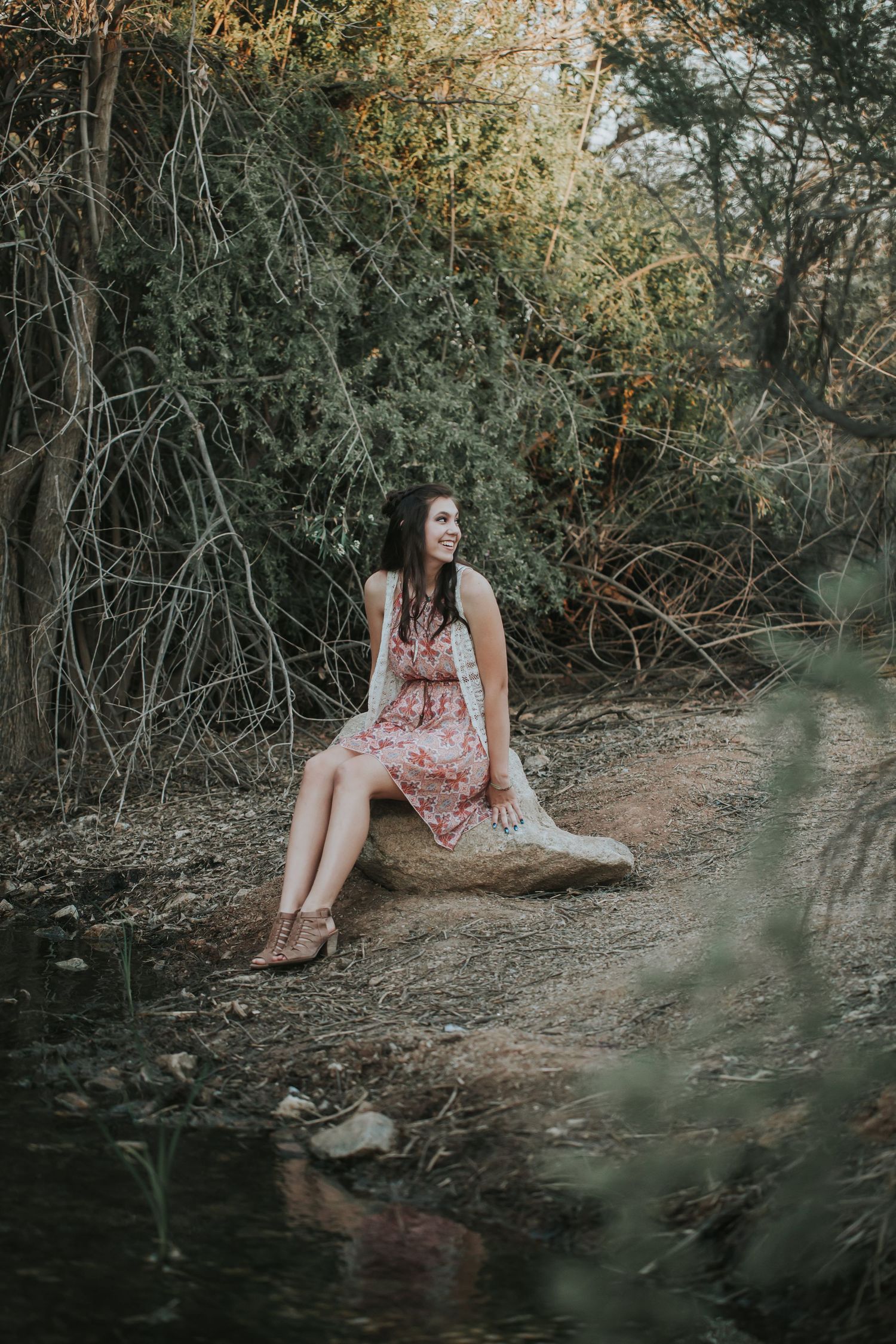 Person in floral dress sitting thoughtfully by a creek in a natural outdoor setting.