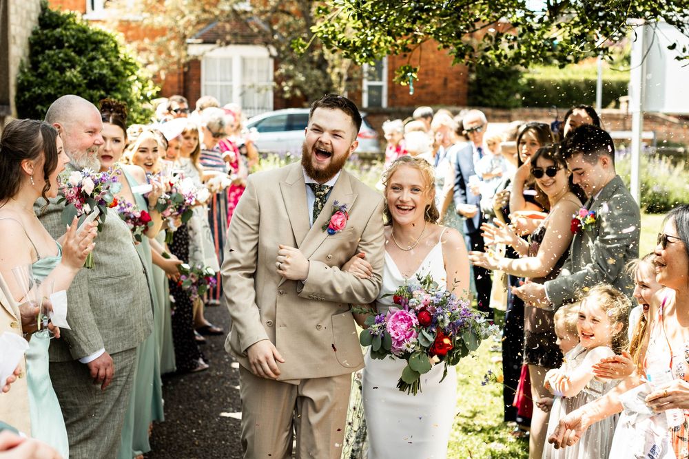 Bride and groom walk through a confetti shower surrounded by bridesmaids and wedding guests outdoors.