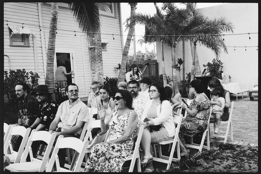 Wedding guests seated in white chairs outdoors at a tropical garden ceremony in black and white.