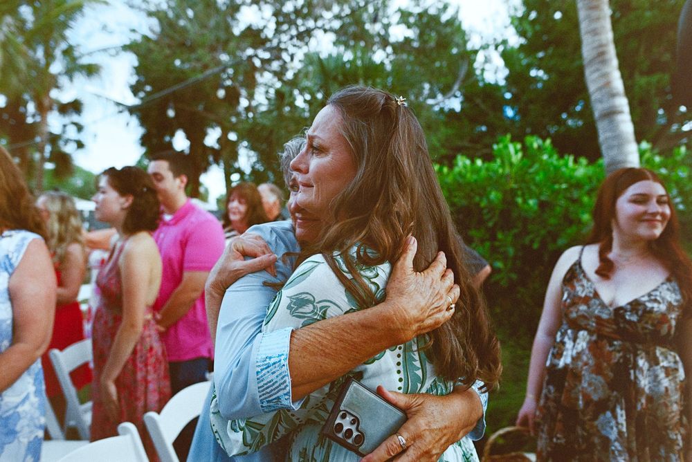 Two women share an emotional hug at an outdoor wedding reception surrounded by palm trees.