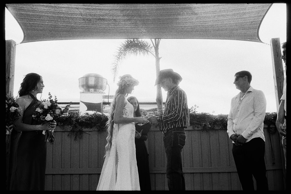 Black and white photo of bride and groom exchanging vows under a canopy with officiant and witness.