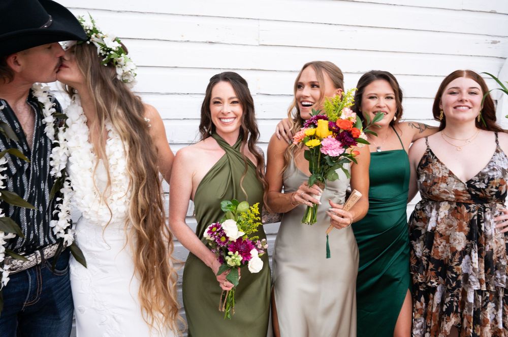 Bride and groom kiss while bridesmaids in green and floral dresses laugh and smile alongside them.