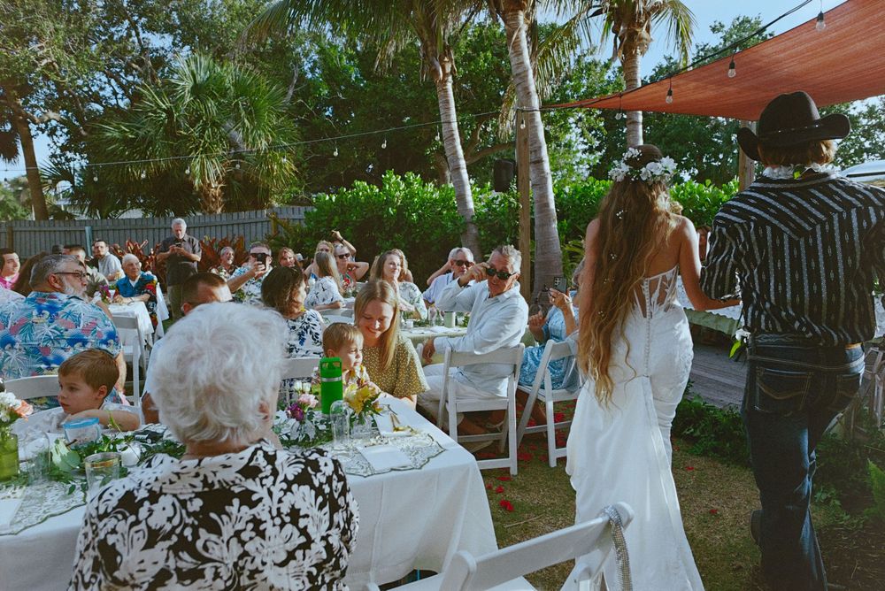 Outdoor wedding reception with guests seated at white tables surrounded by palm trees.