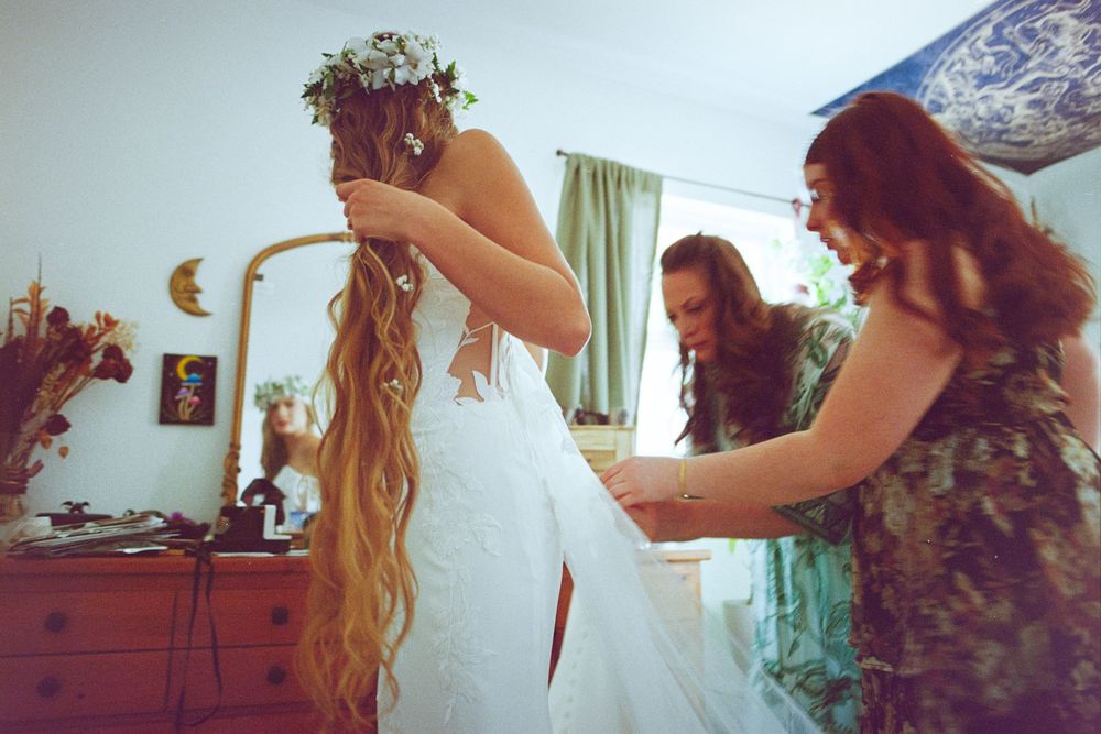 Bride with long hair and floral crown gets dress laced up by two bridesmaids in a warmly lit bedroom.