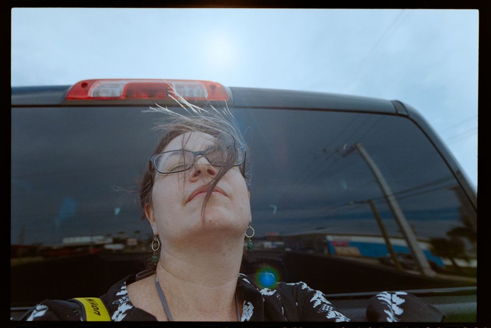 Woman with glasses tilts head back near a truck under a bright sky on a cloudy day.