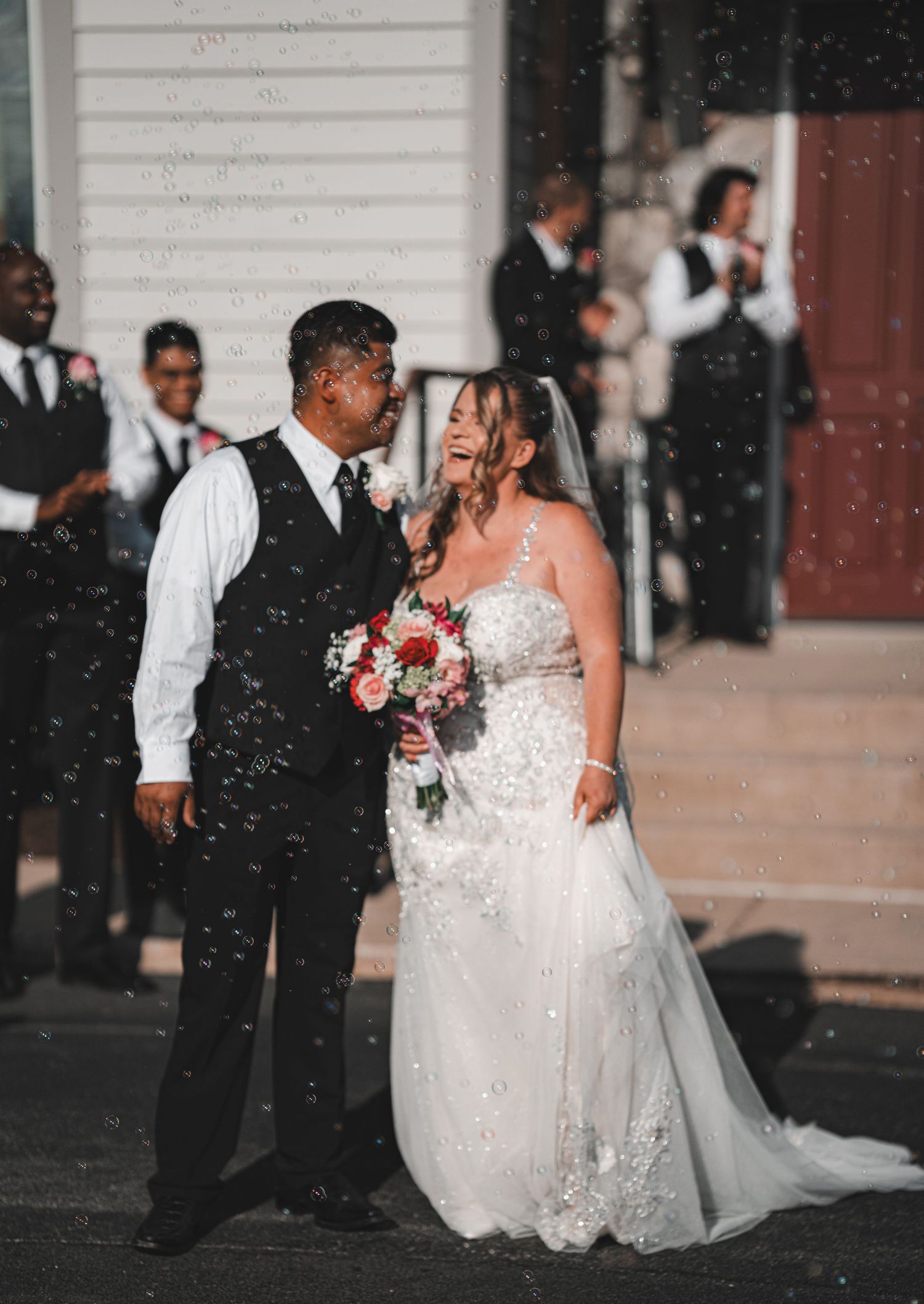 Bride and groom smiling together outside a church surrounded by bubbles and wedding party.