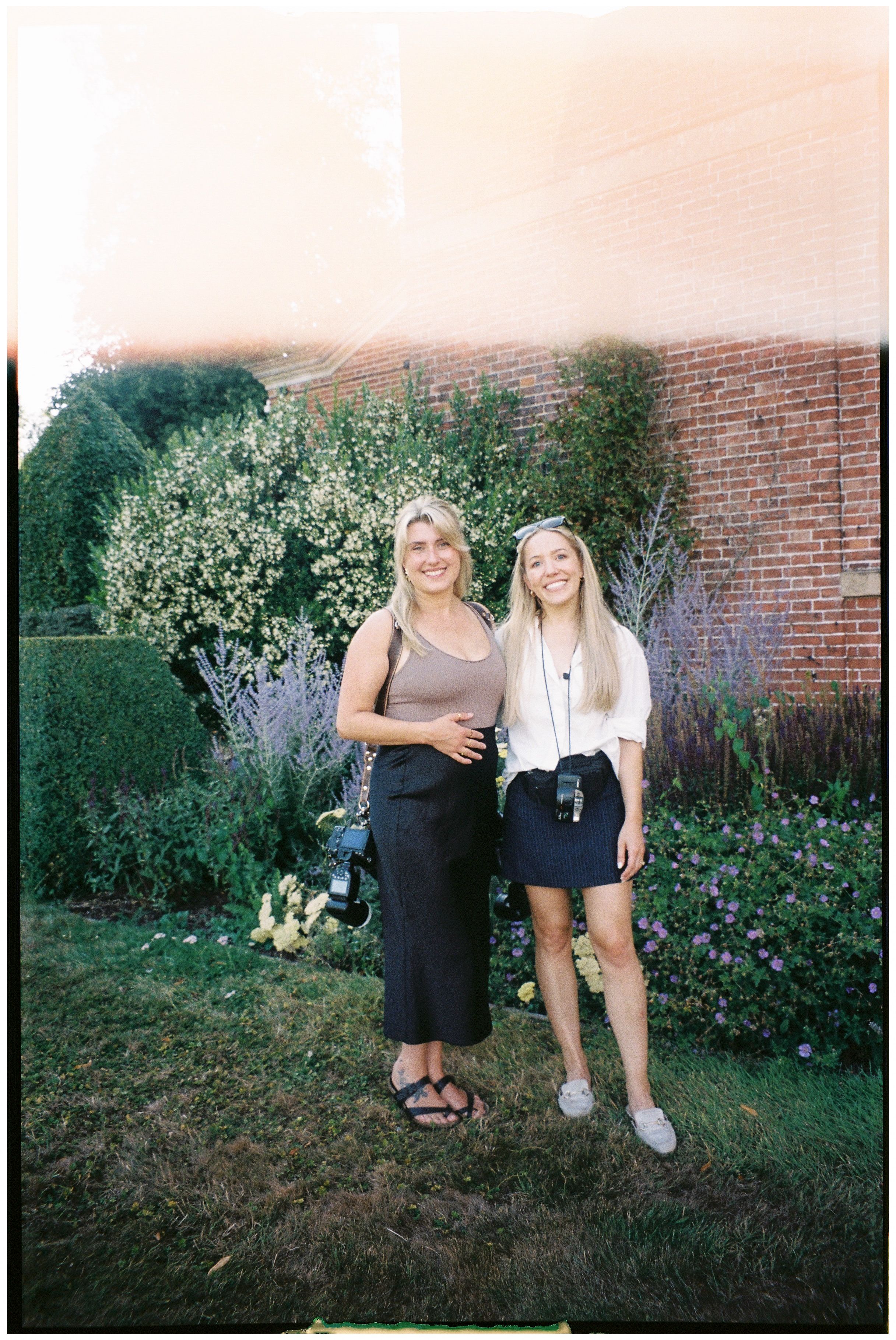 Two friends pose together in a garden with flowering plants and a brick wall backdrop during golden hour.