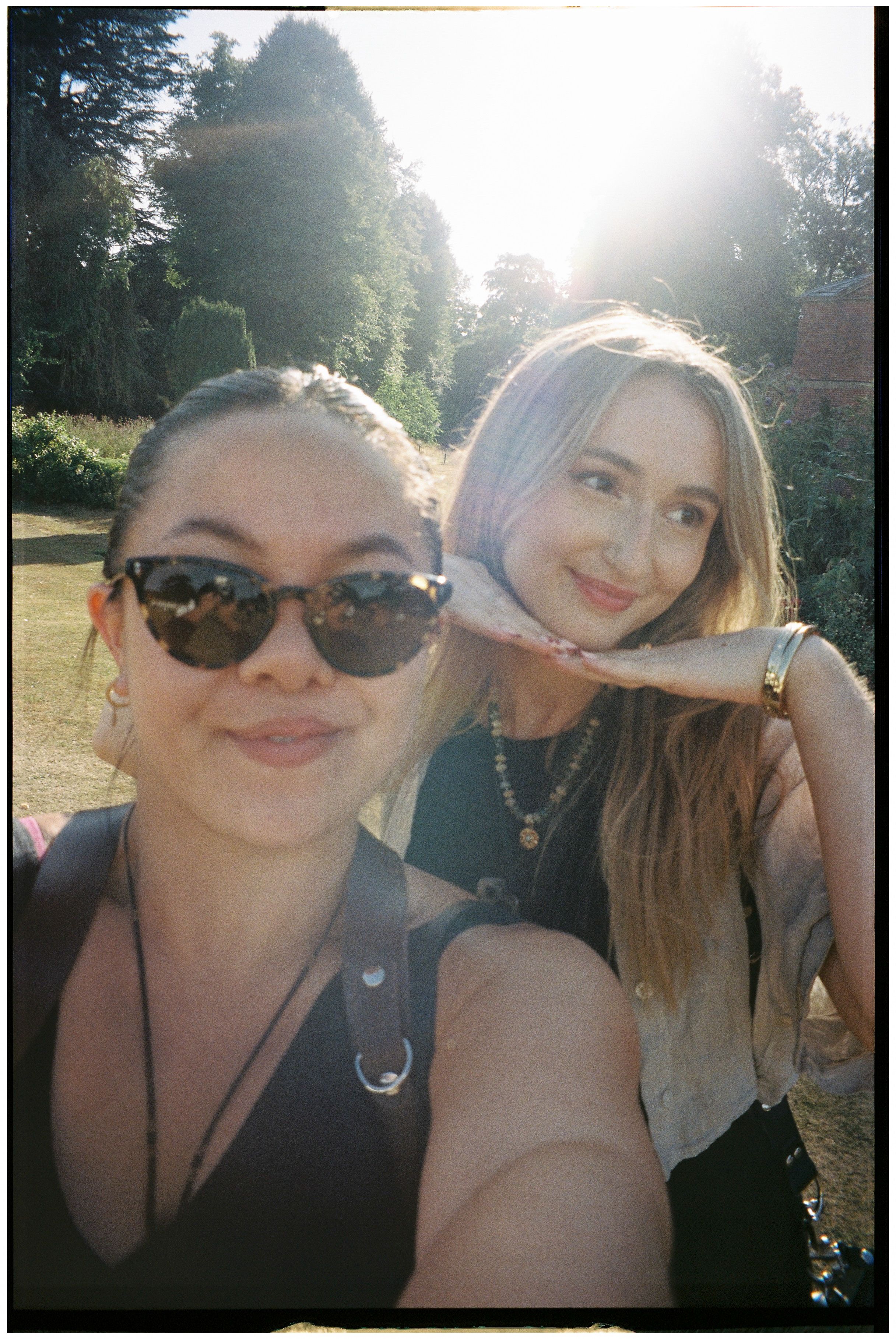 Two friends pose together outdoors on a sunny summer day with trees in the background.