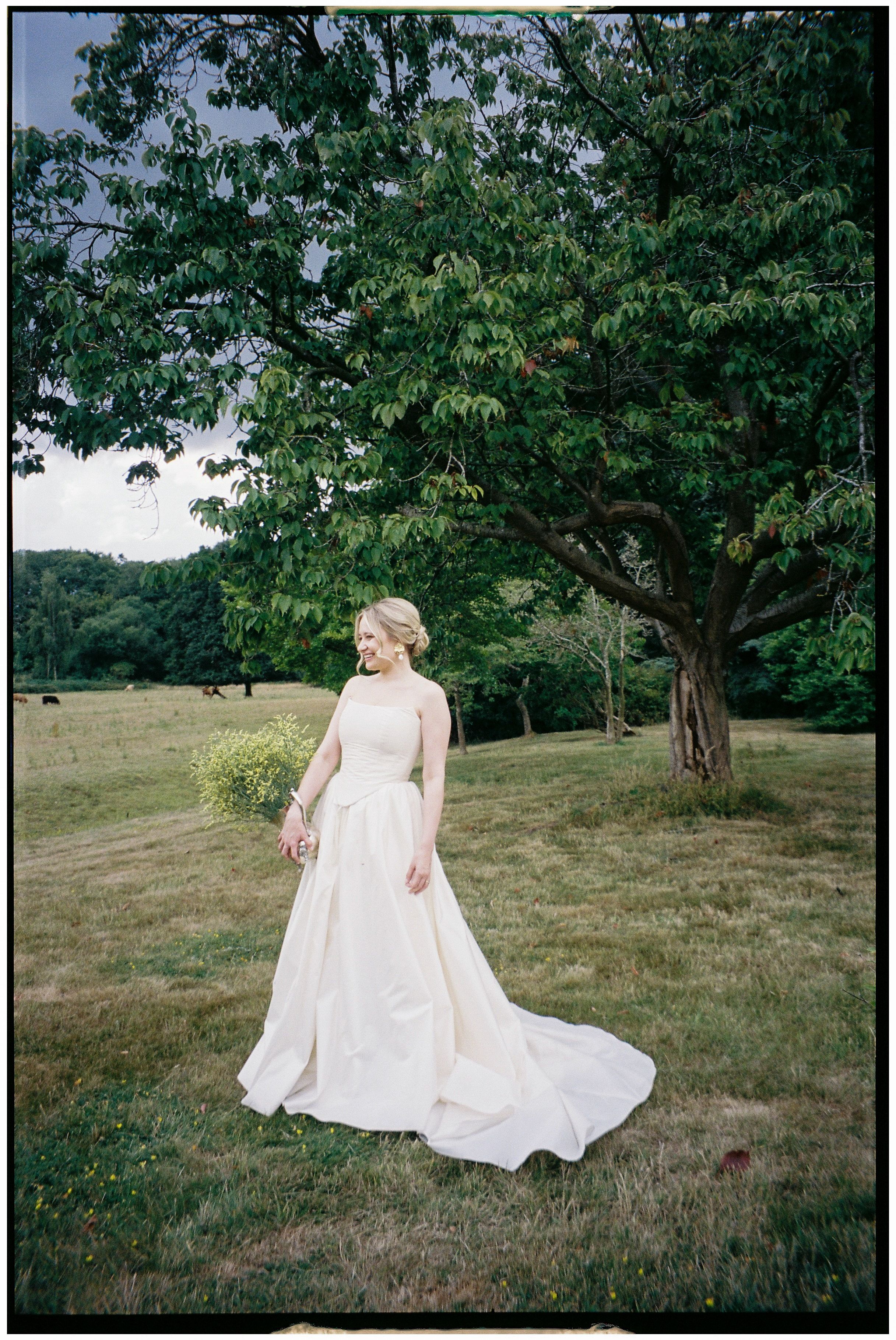 A person in a flowing white wedding dress walks through a serene meadow surrounded by lush green trees.