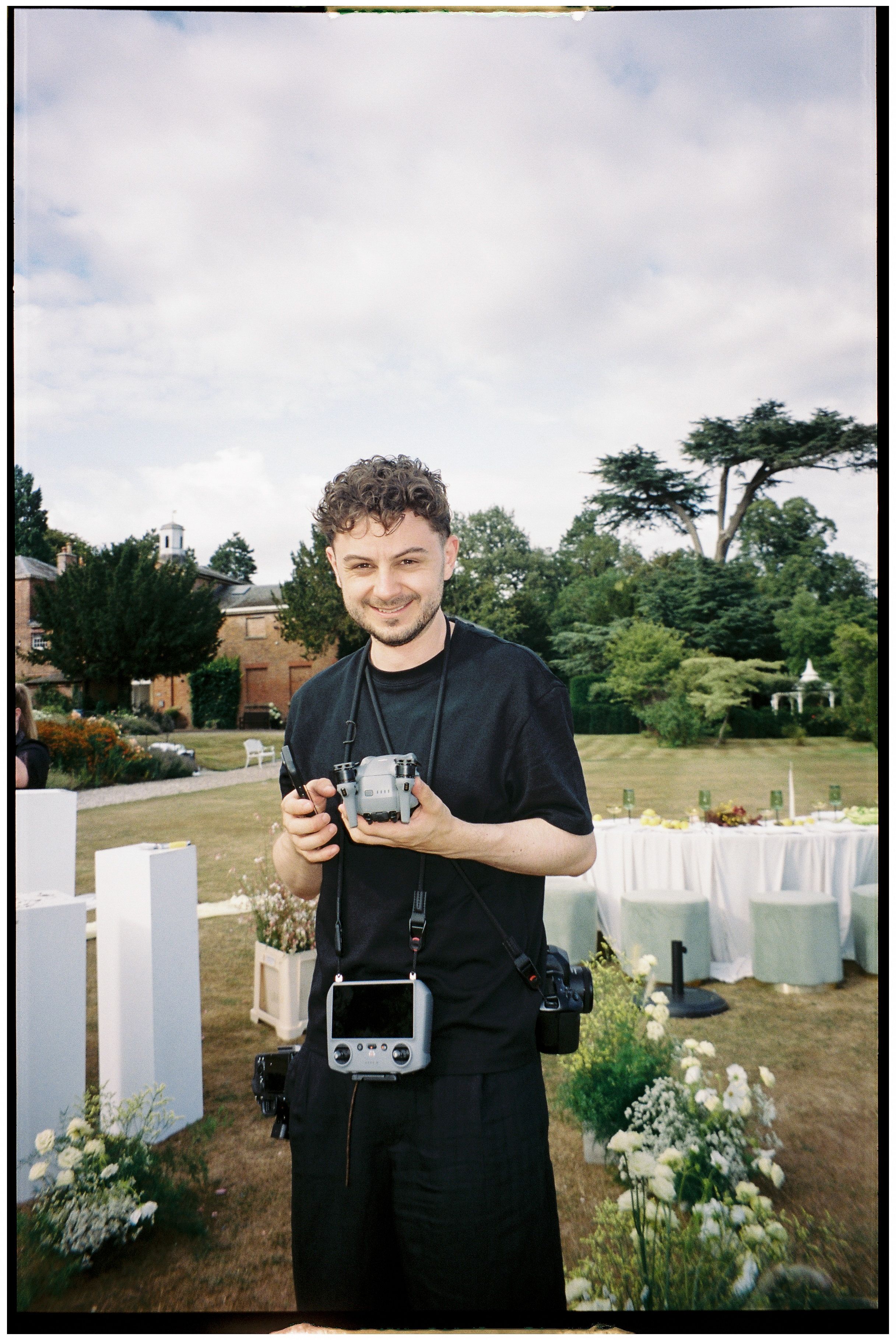 A photographer in black attire holds vintage cameras while standing in a garden with white chairs set up outdoors.