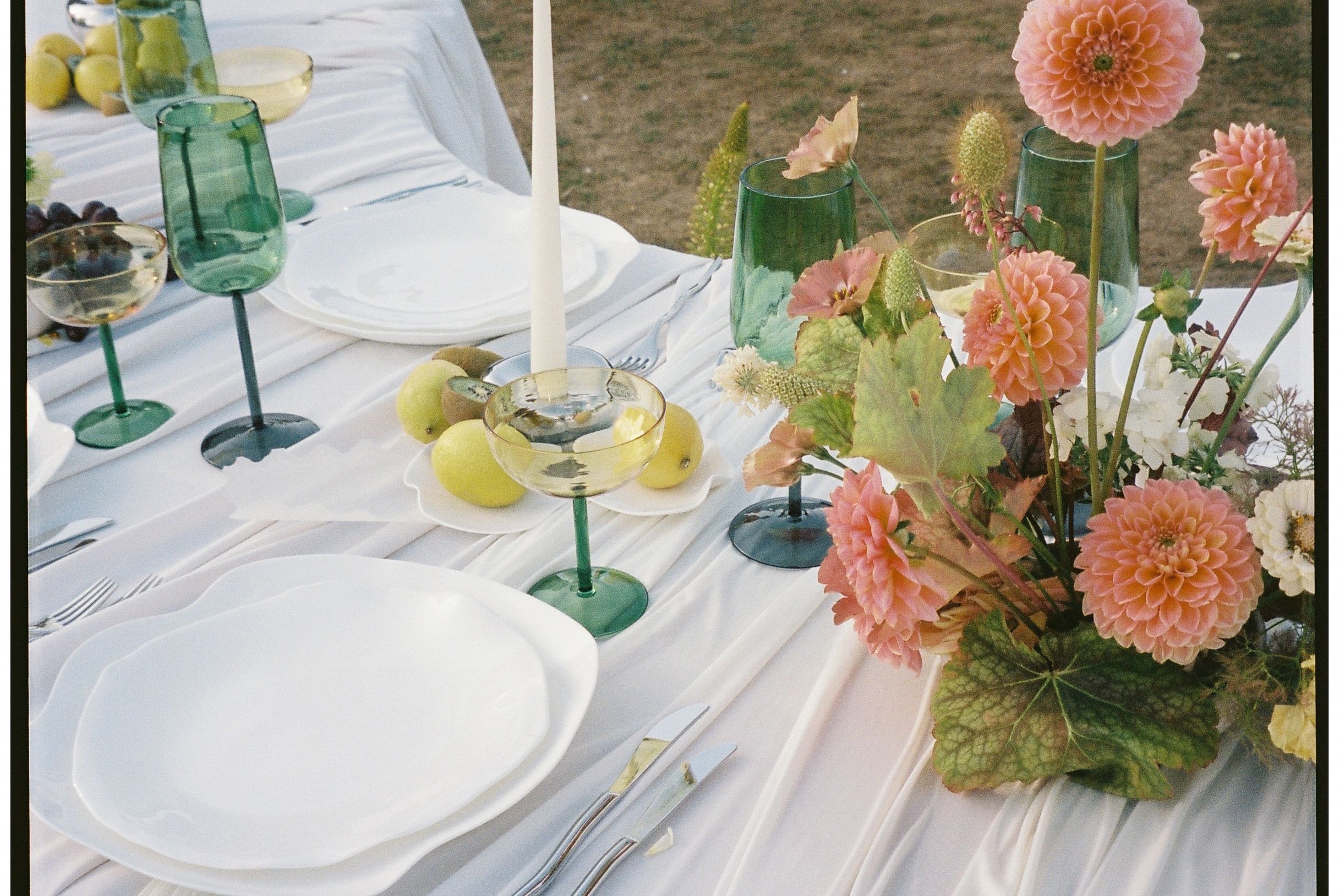 Elegant table setting with coral dahlias, green glassware, and white linens for an outdoor garden party.