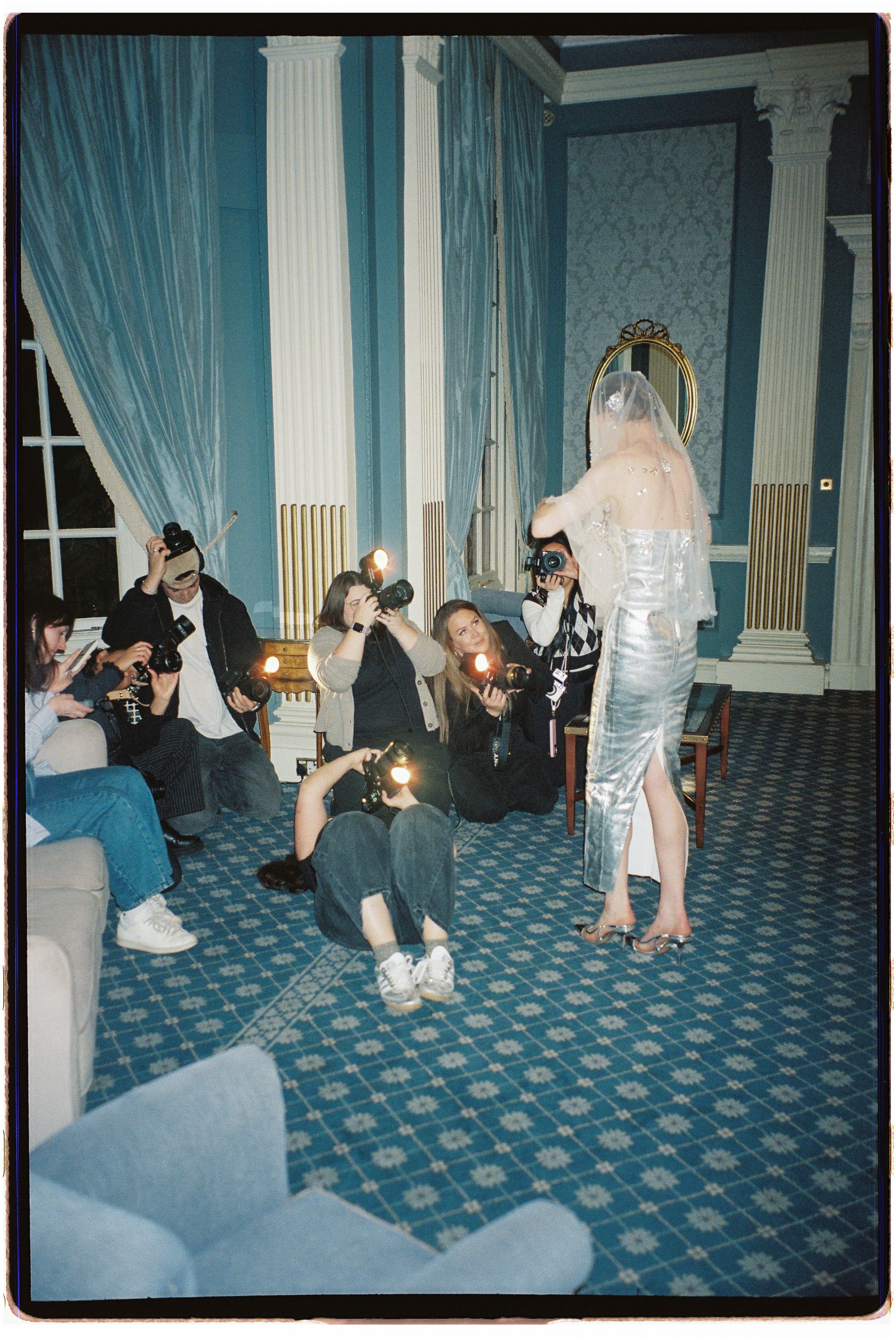 Party guests in casual and formal attire gather on a blue patterned carpet in an elegant room with teal curtains and columns.