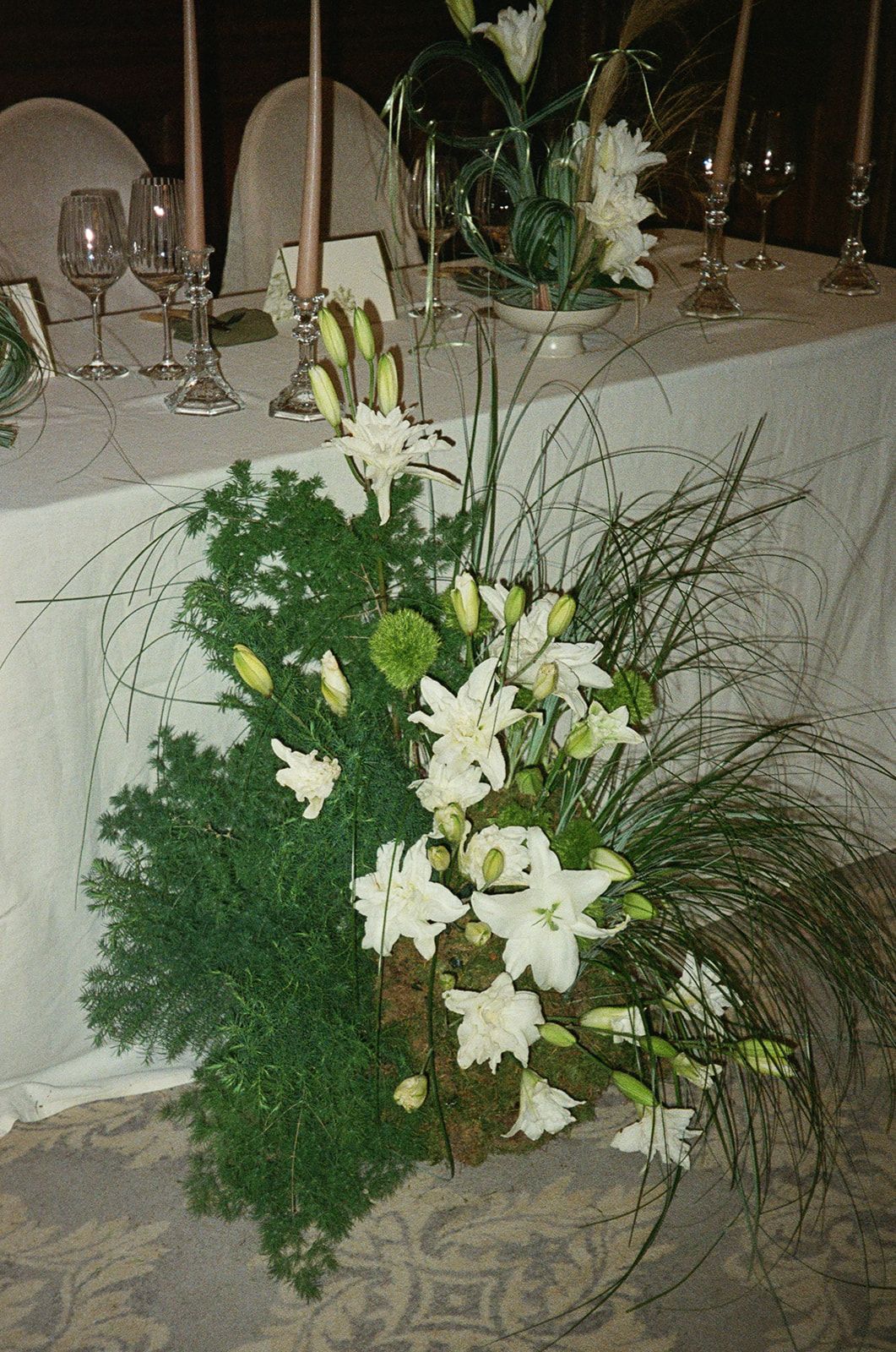 Elegant white lily floral centerpiece with lush greenery on a formal dining table with white tablecloth and wine glasses.
