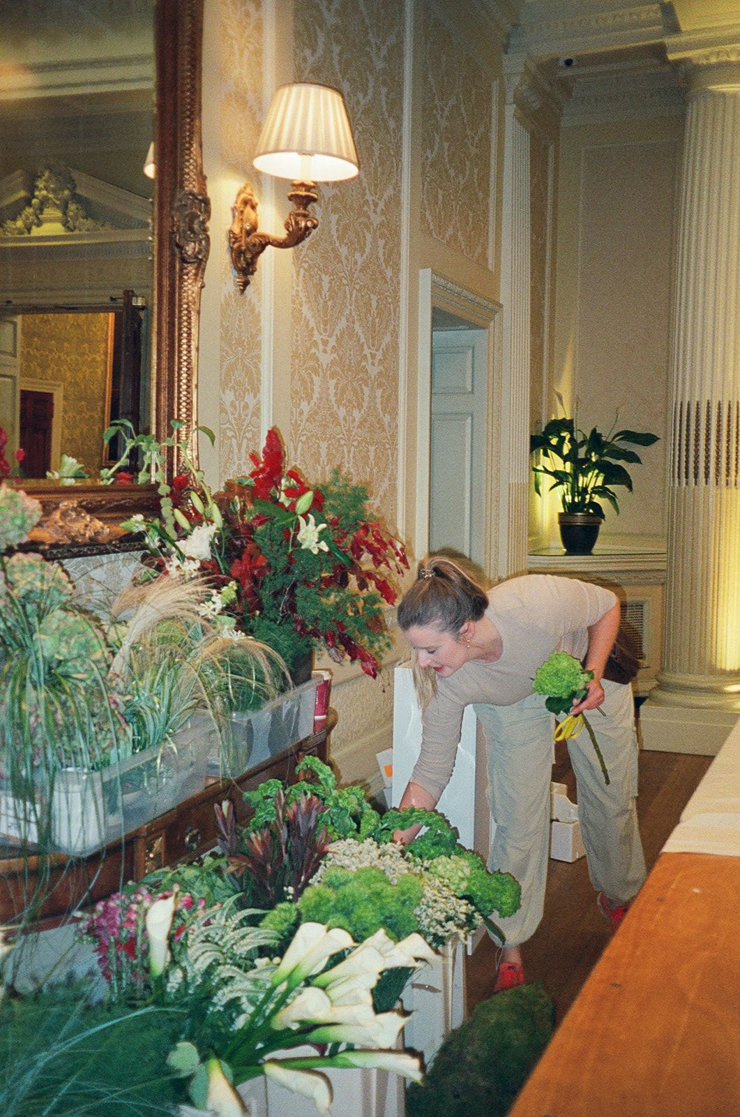 Floral designers arrange elaborate green and white flower displays along a long table in an elegant room with ornate wallpaper.
