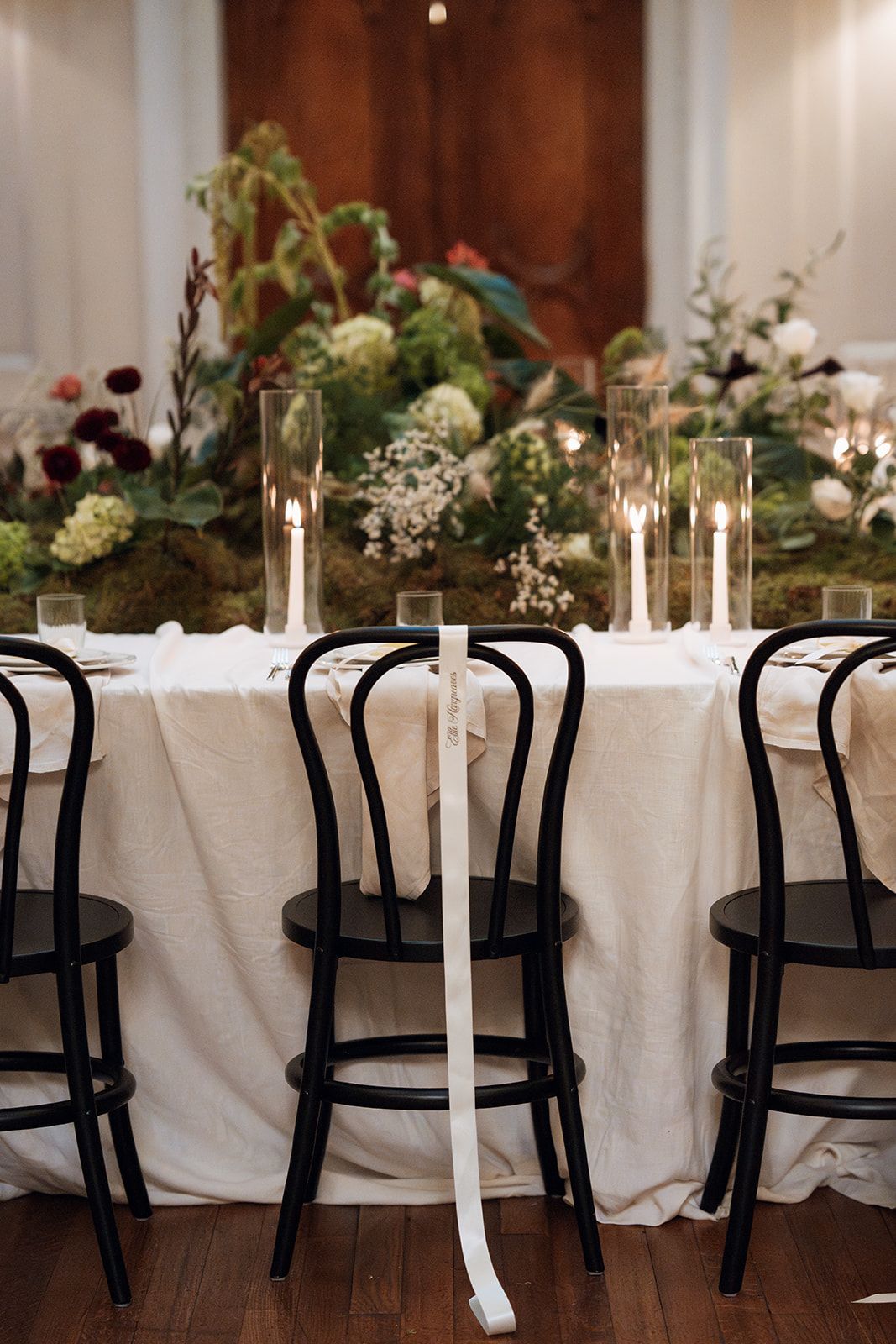 Elegant wedding reception table with white linens, black chairs, and delicate floral centerpieces illuminated by candlelight.