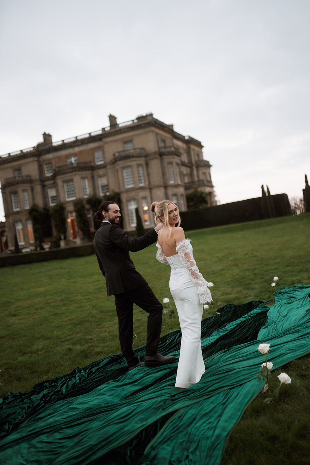 Wedding couple playfully slides down a green slope in front of a grand manor house on an overcast day.
