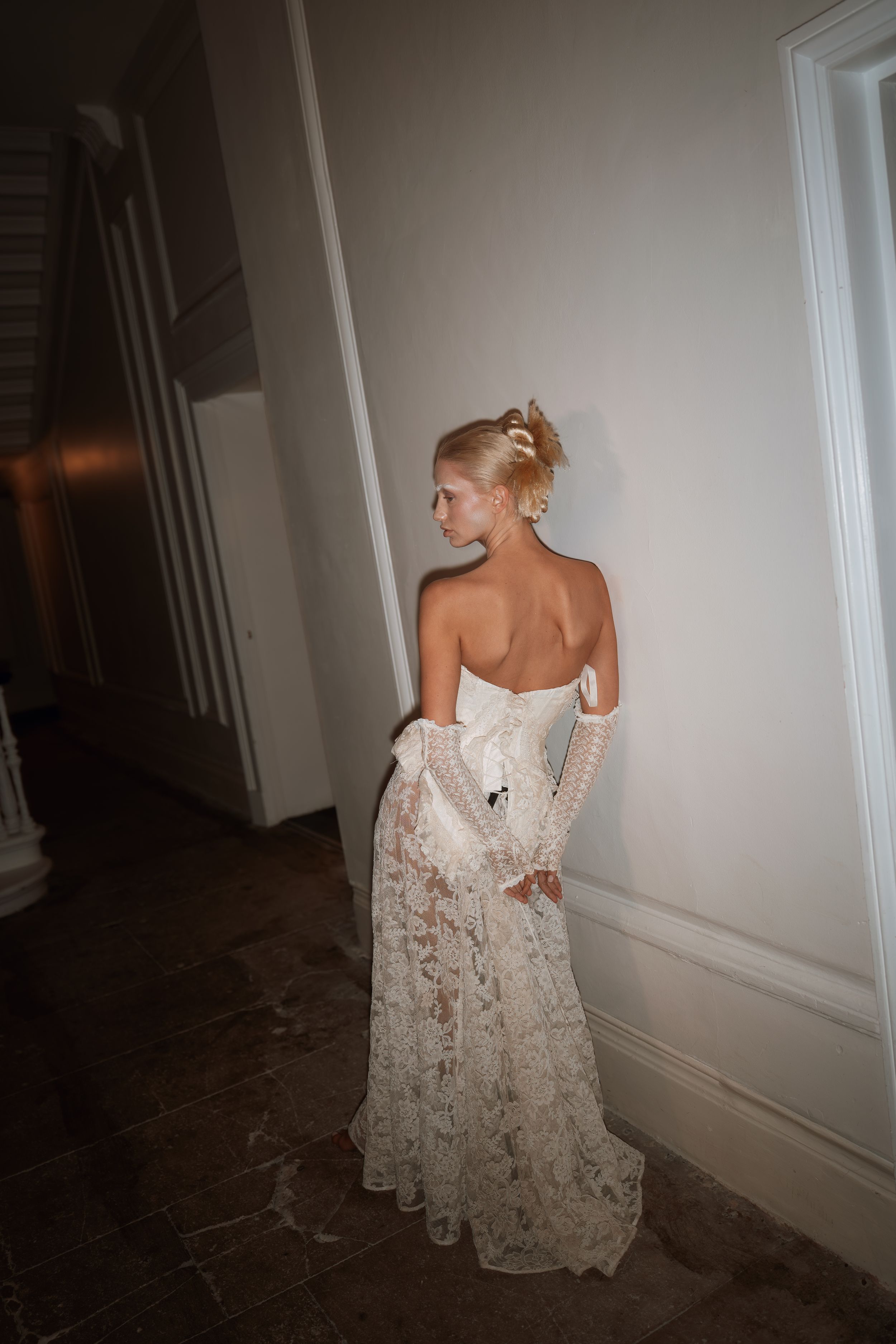 A bride in a strapless lace wedding gown stands with her back turned in a dimly lit hallway.
