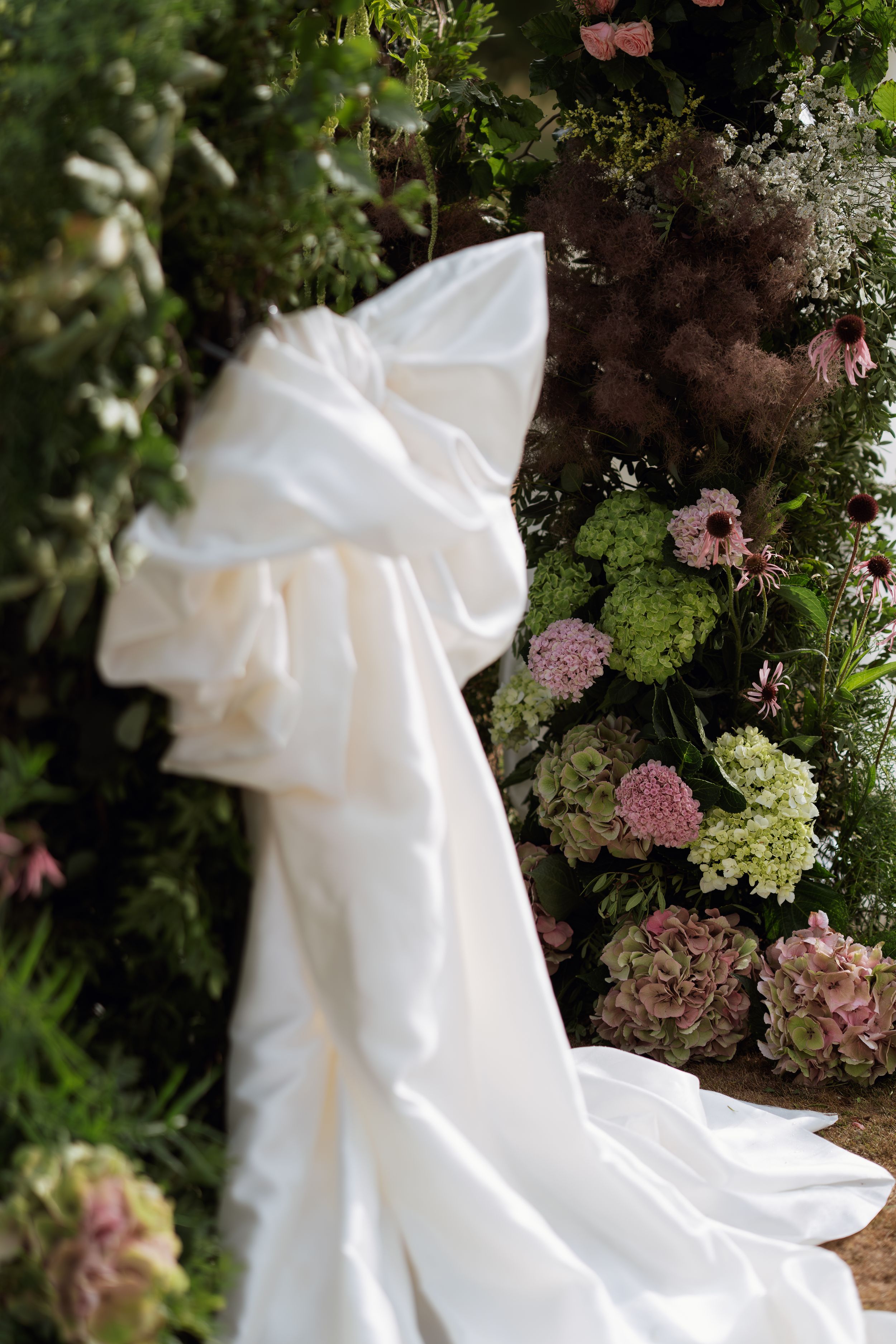 A flowing white wedding dress with a dramatic train is displayed against a backdrop of pink flowers and green foliage.