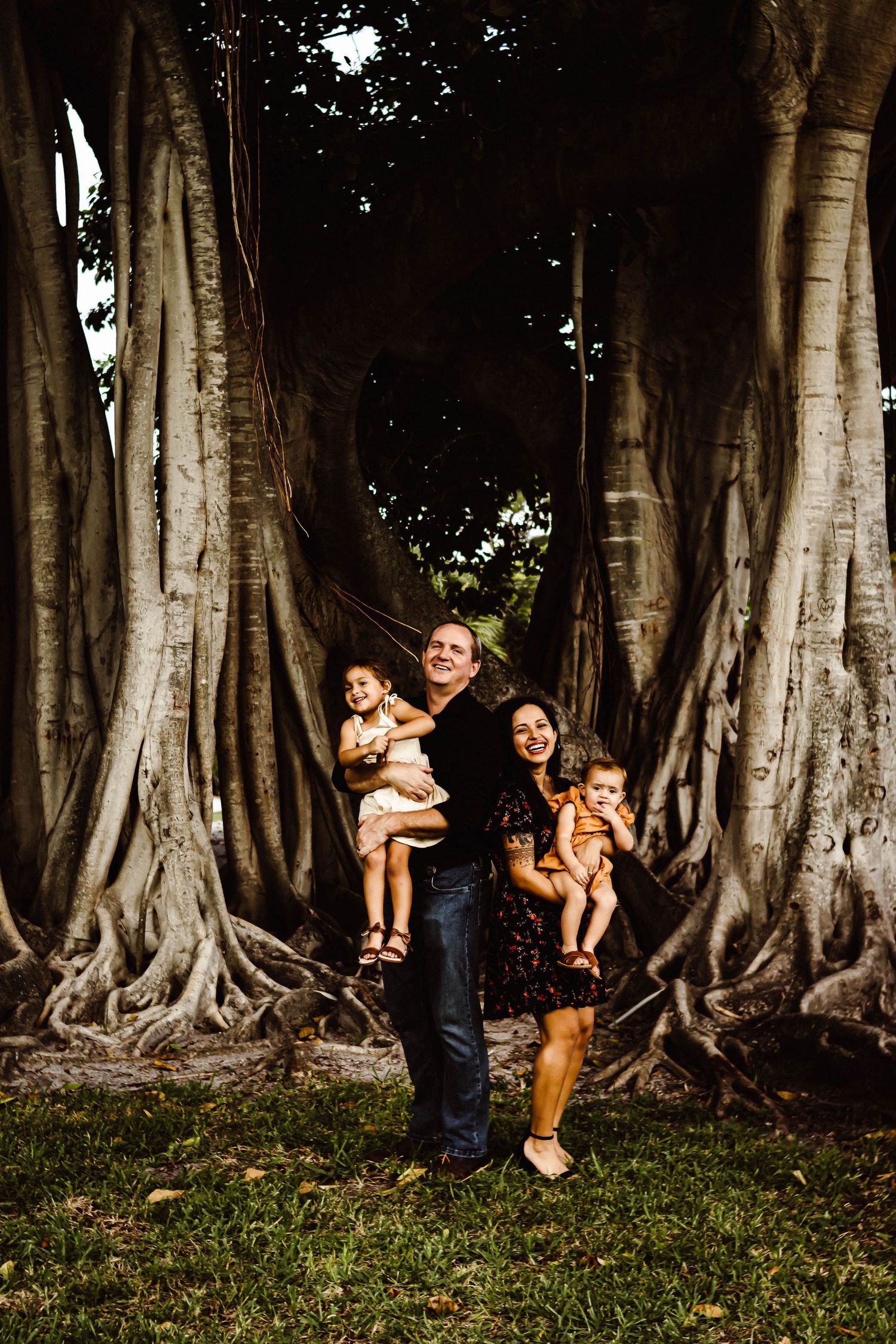 Family poses together between massive banyan tree trunks in a tropical garden setting at sunset.