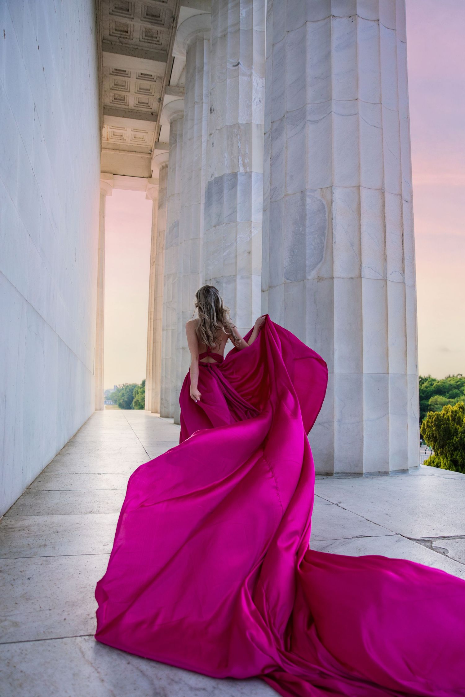 A flowing magenta gown with long train billows dramatically between classical white marble columns at sunset.
