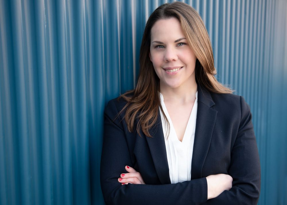 Professional business headshot of someone in a black blazer and white blouse against a blue background.