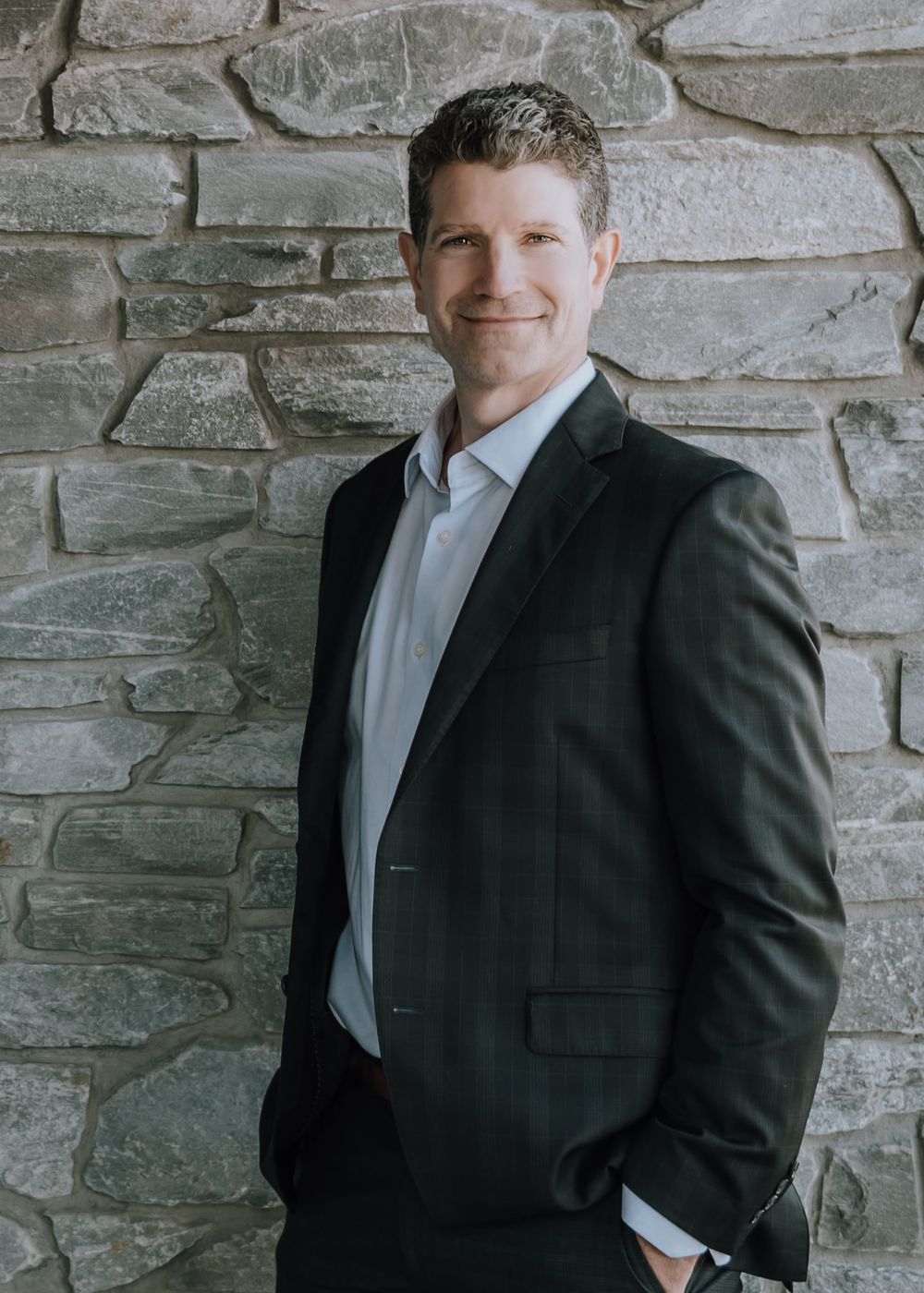 Professional portrait of a person in a dark suit and white tie against a rustic stone wall background.
