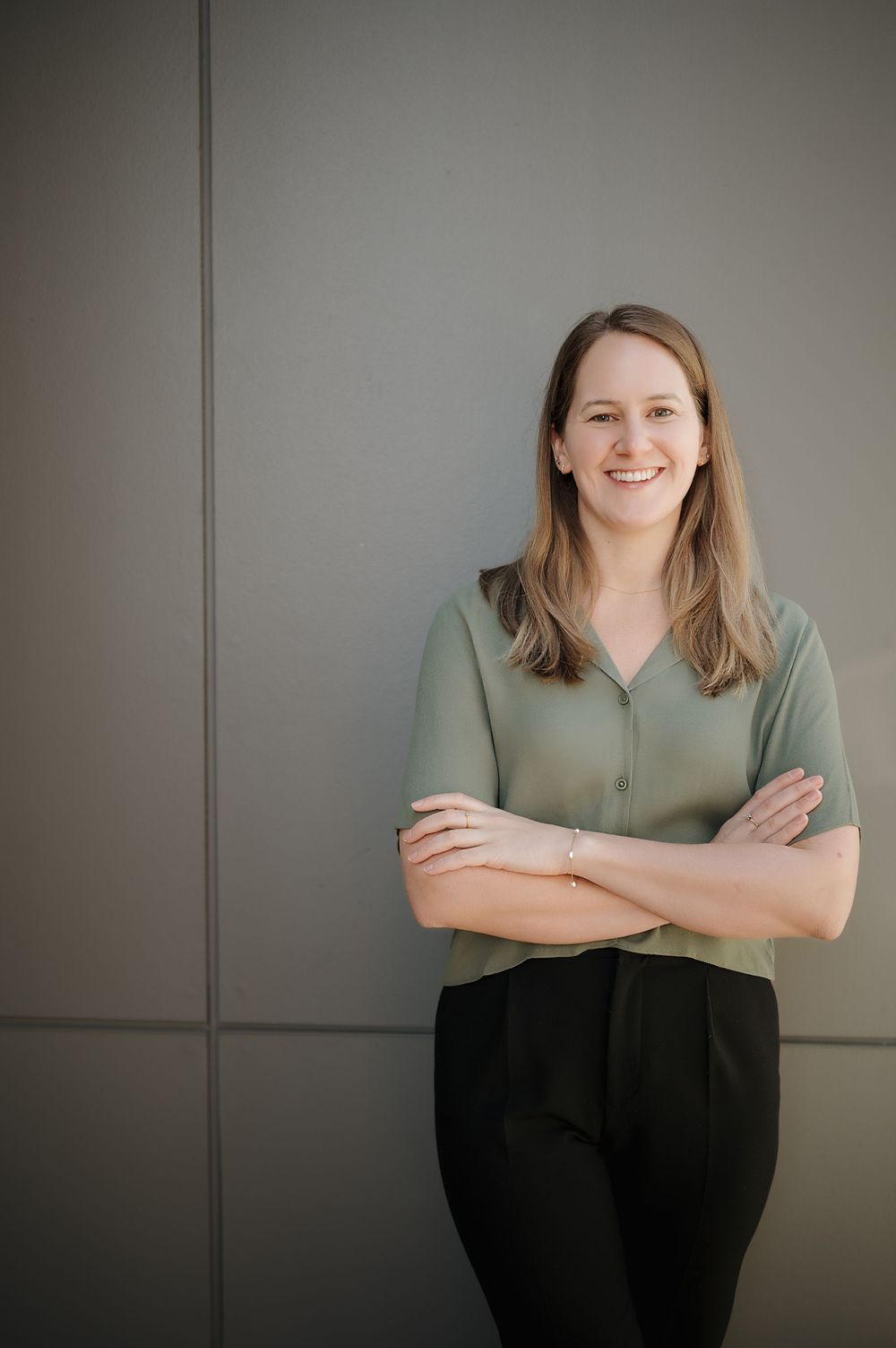 Professional portrait of someone in a sage green blouse and black pants standing with arms crossed against a gray wall.