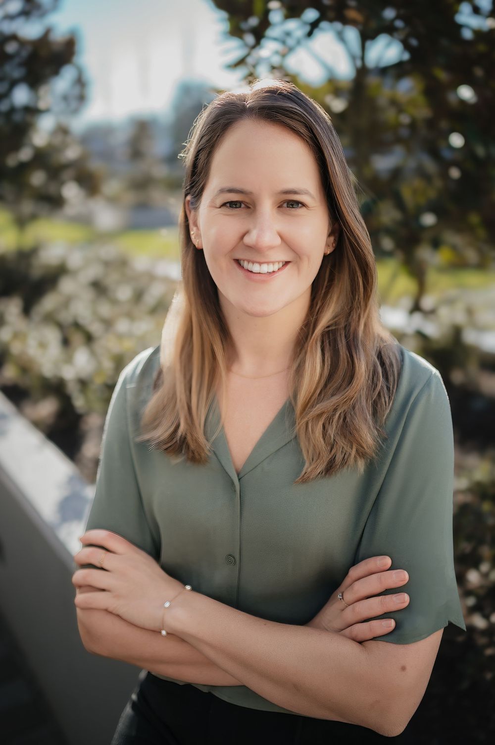 Professional headshot of someone in a sage green button-up top against a blurred natural outdoor background.