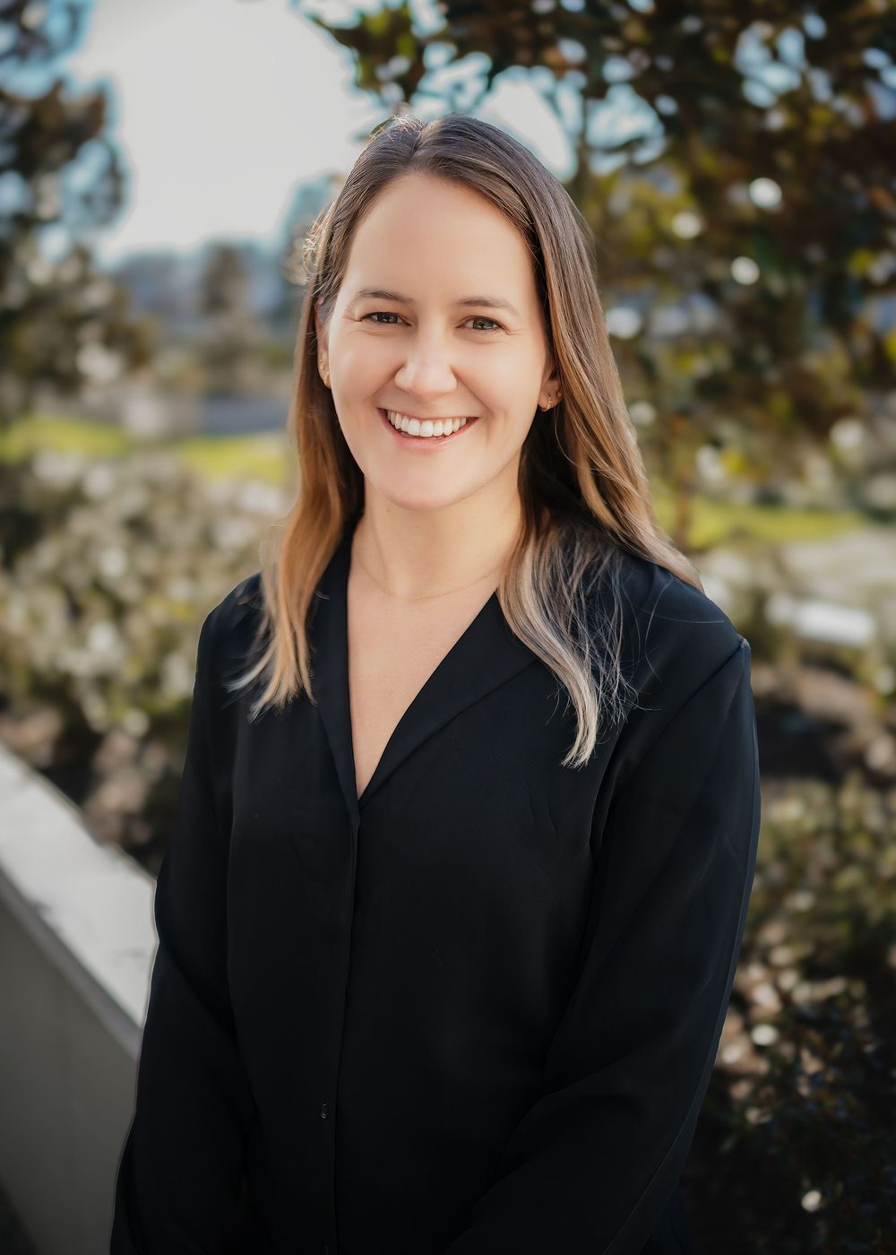 Professional portrait of someone in a black blazer smiling warmly against a blurred outdoor background.