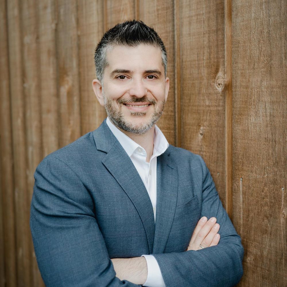 Professional headshot of someone in a gray suit jacket against a wooden fence background.