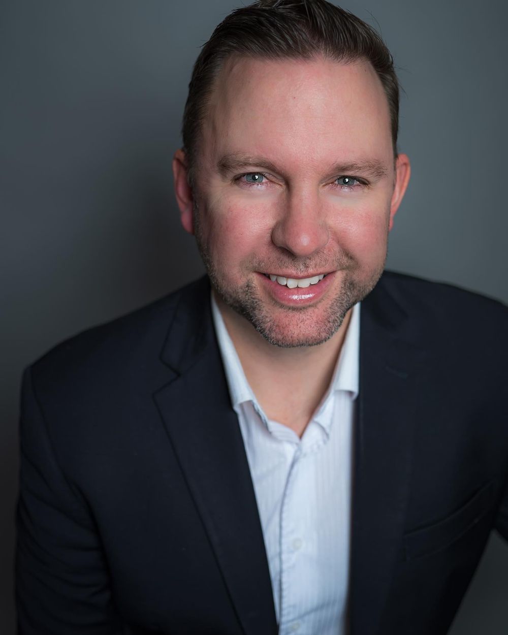 Professional headshot of a smiling person in a dark suit jacket and white shirt against a gray background.