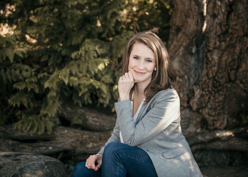 Woman in gray sweater sits outdoors on fallen log while smiling at camera in autumn forest setting.