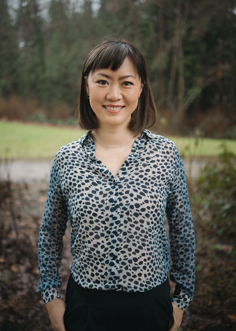 A woman in a leopard print top and black skirt poses outdoors against a wooded background.