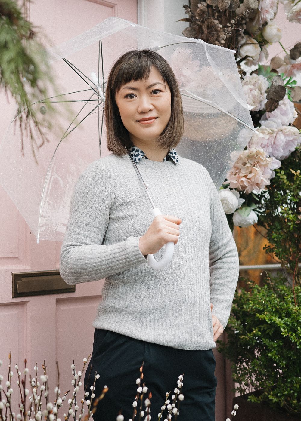 A person in a gray sweater and black skirt poses against a pink wall with cherry blossoms and greenery.