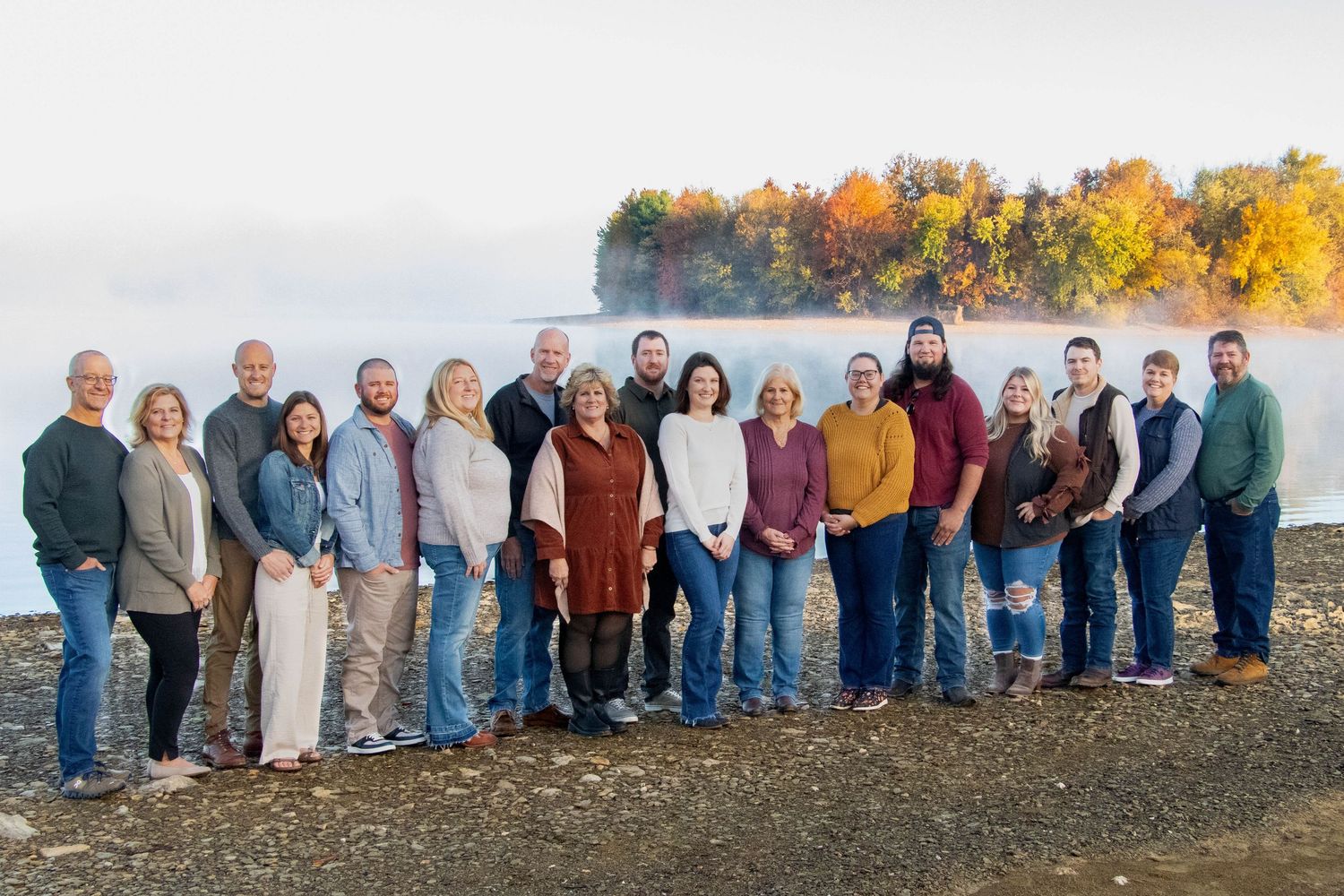 Large extended family group portrait on rocky lakeshore surrounded by colorful fall foliage.