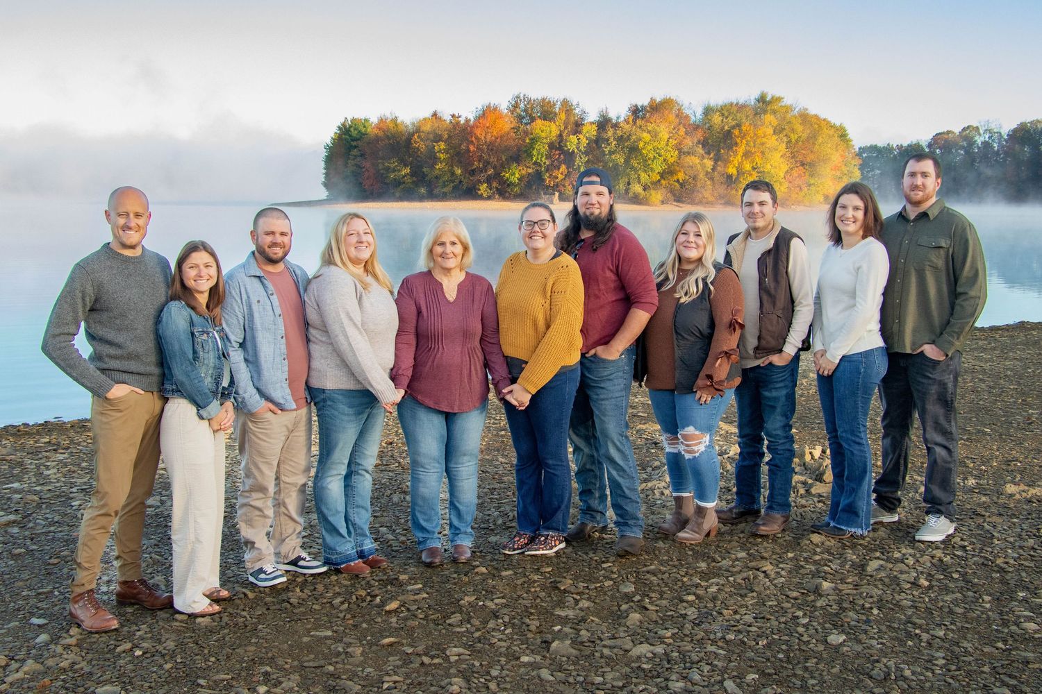 Group of ten adults smiling together on a rocky lakeshore with misty autumn lake backdrop.
