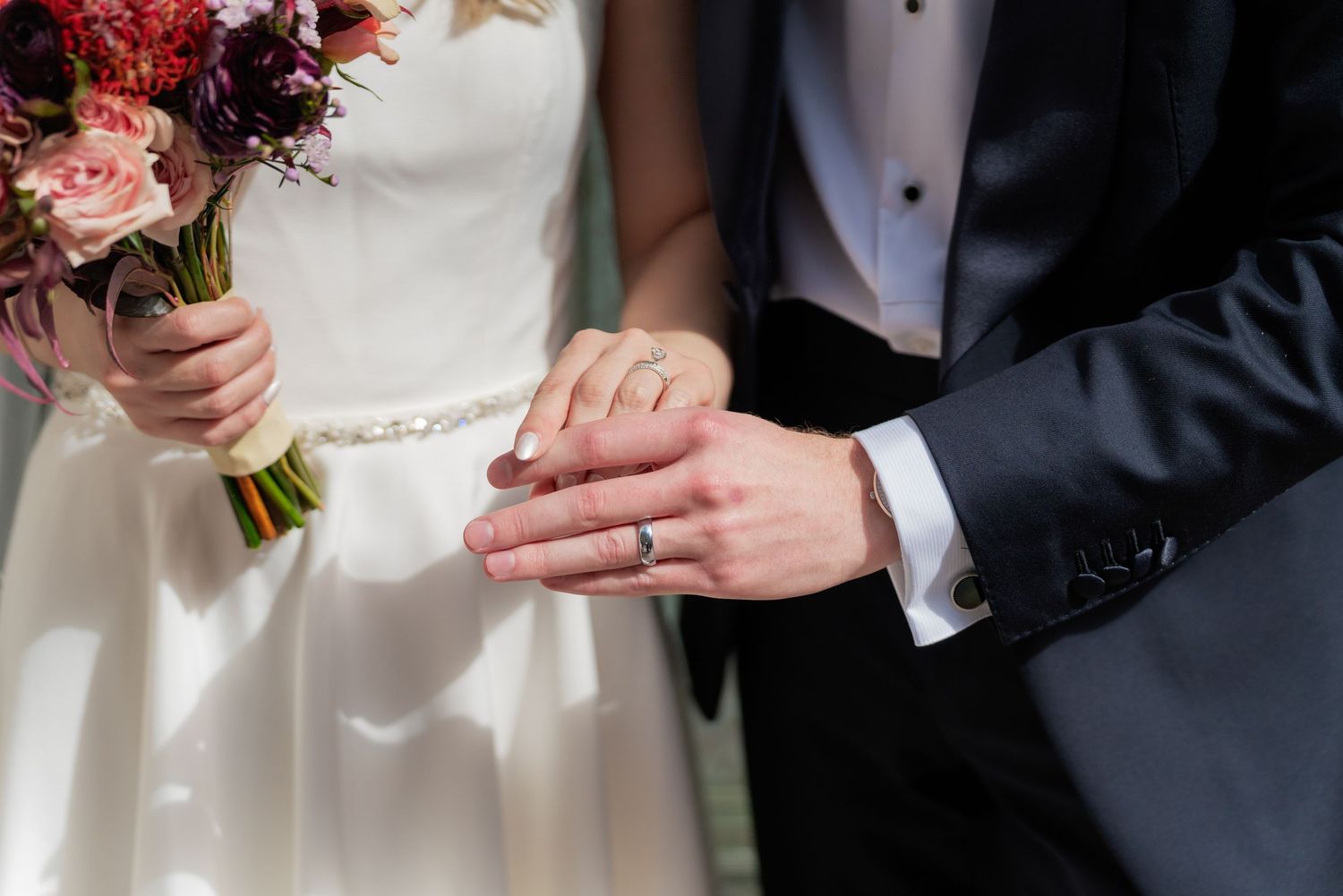 A couple displays their wedding rings while holding a pink and white floral bouquet.
