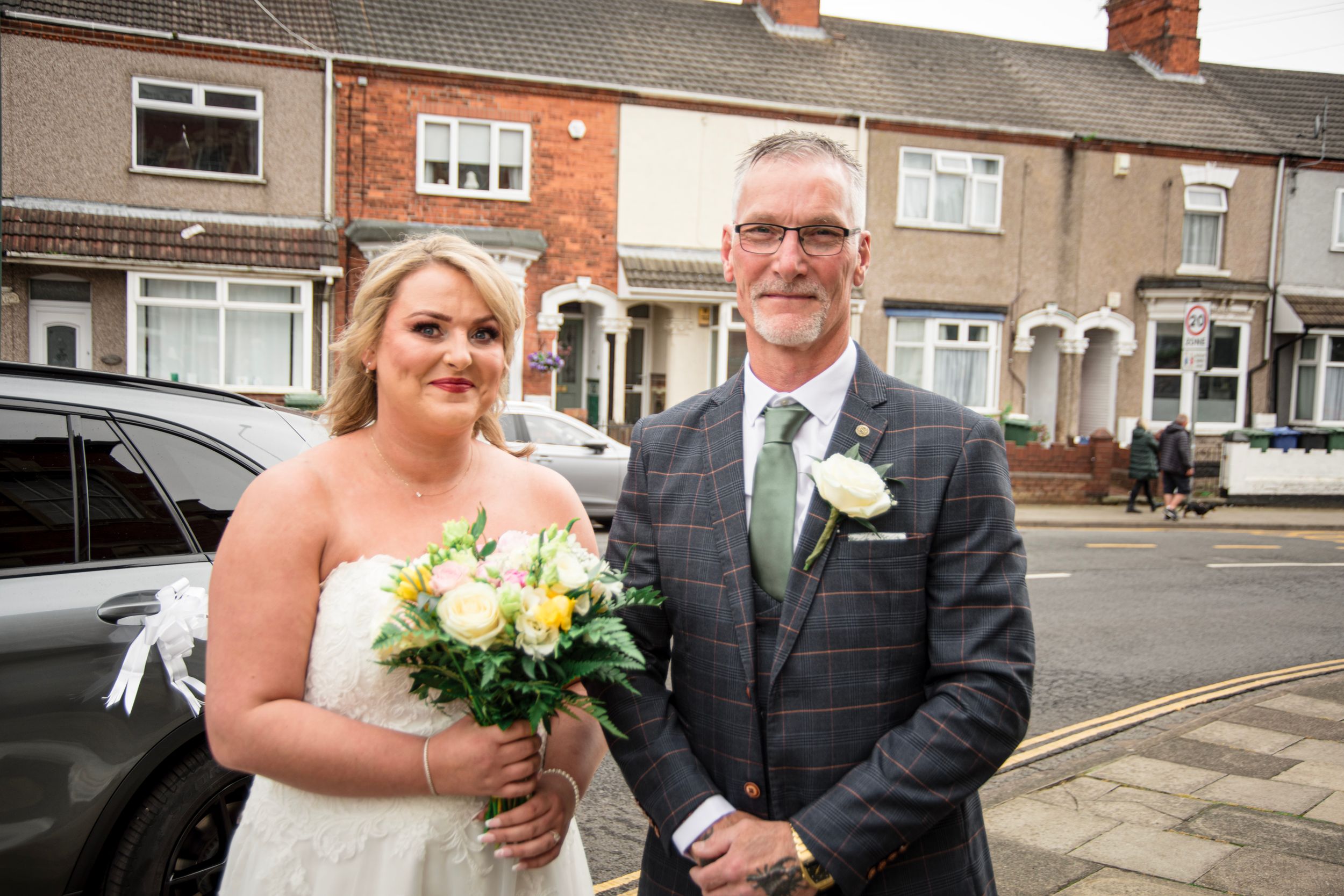 A wedding couple poses together on a residential street with brick houses in the background.